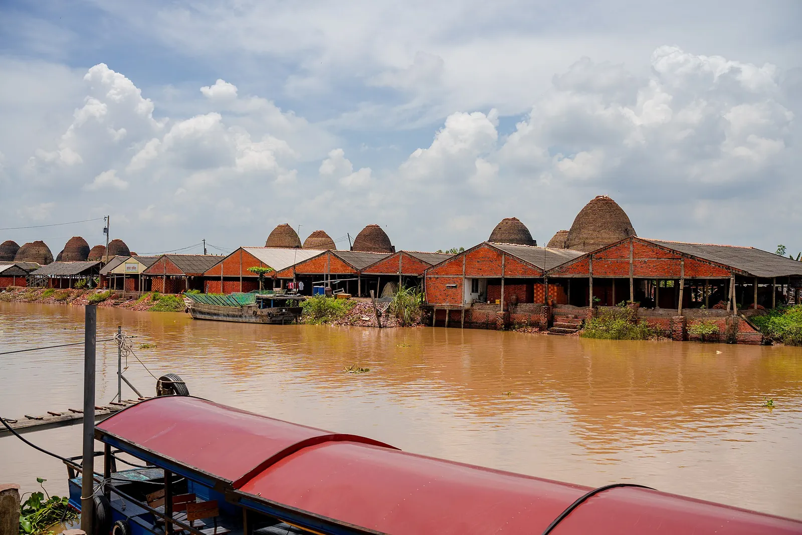 A row of domed red-brick kilns with tiled roofs along a wide brown waterway at Mang Thit, with a covered boat in the foreground