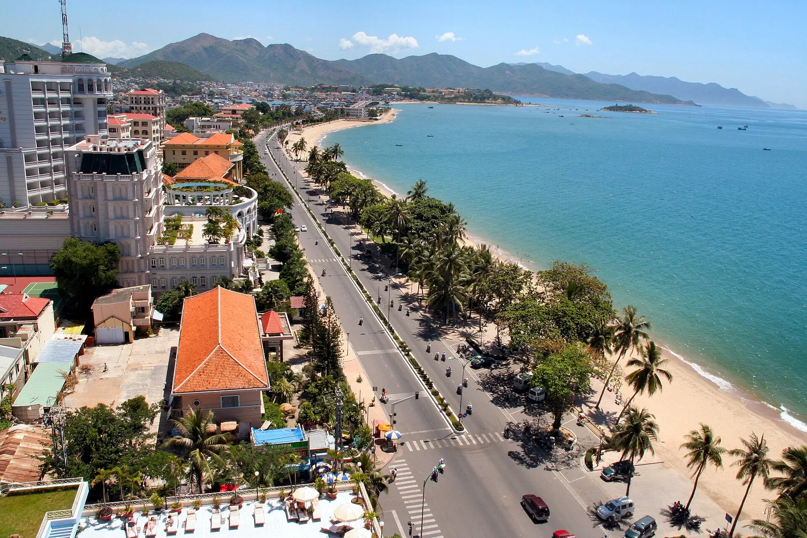En palmebeplantet kystvej, der snor sig langs en sandstrand og det blå hav ved Nha Trang, med hoteller langs vejen
